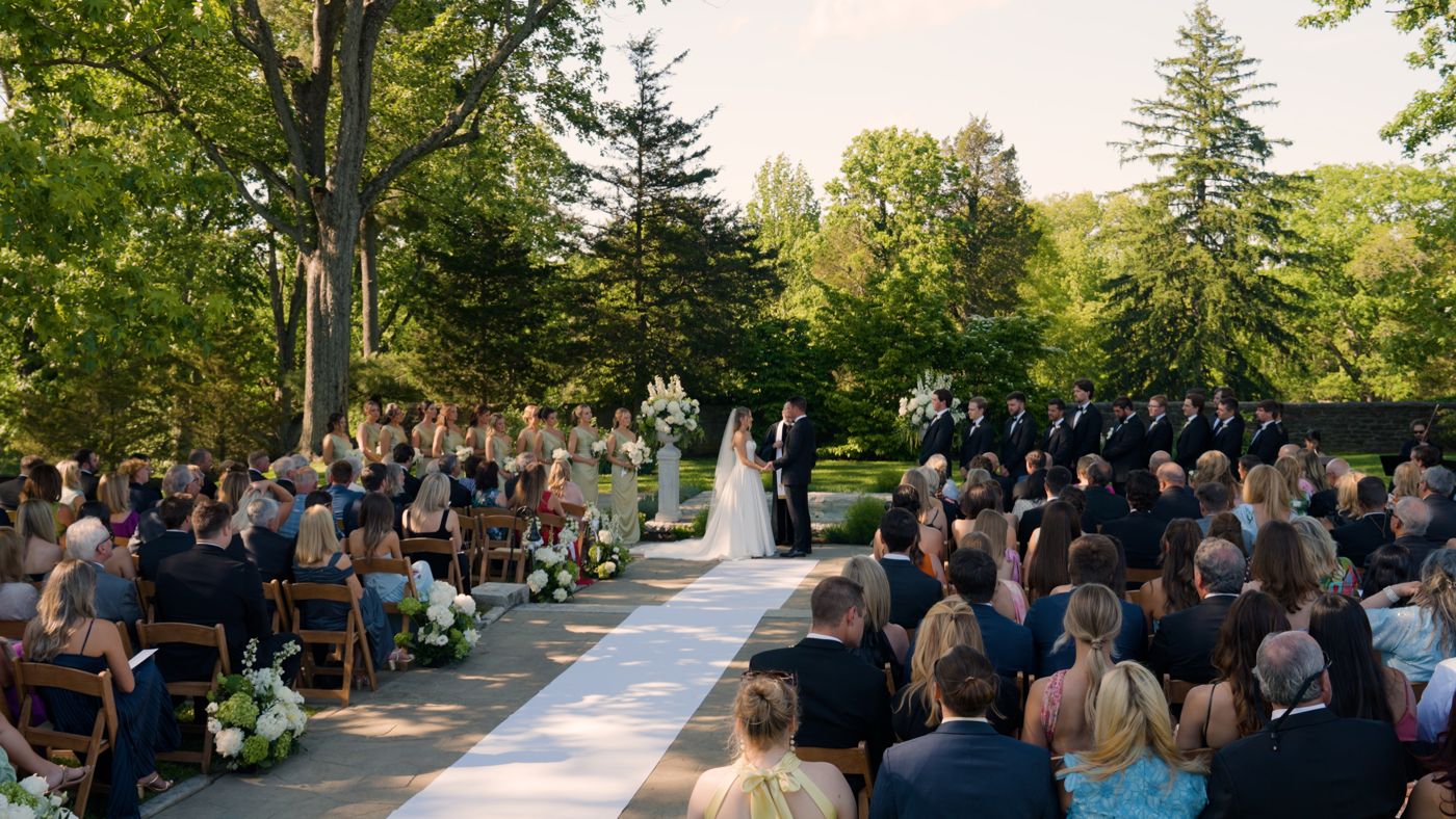 Wide shot of an outdoor garden wedding ceremony with guests seated along a white aisle runner